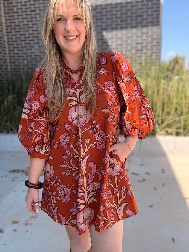 A woman wearing a rust-orange short-sleeve dress with a pink and beige floral pattern, standing outdoors in front of a brick wall.