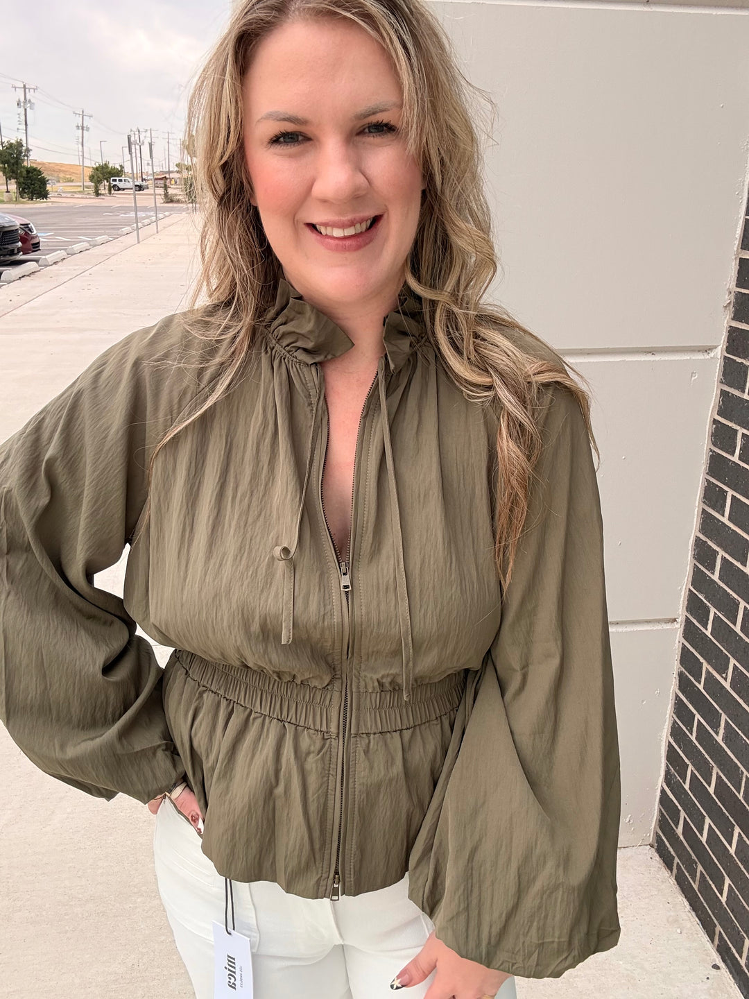 A woman wearing an olive green crinkled zip-up jacket with a high gathered collar and long balloon sleeves, paired with white pants, standing outdoors next to a light-colored wall.