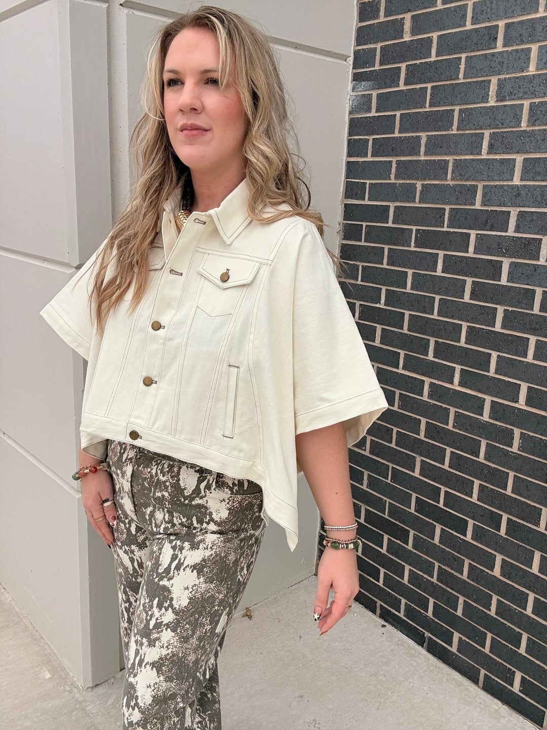 A woman wearing a cream-colored short-sleeve cropped denim jacket and gray-and-white patterned pants, standing outdoors next to a gray brick wall.