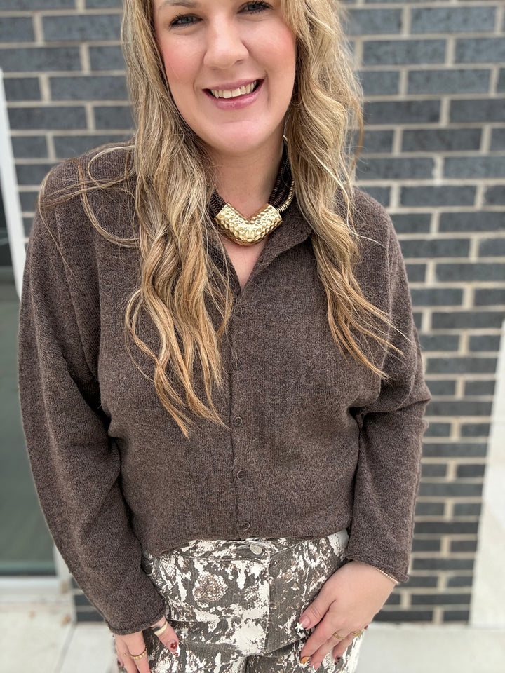 A woman wearing a brown long-sleeve button-up shirt and a chunky gold statement necklace, standing in front of a gray brick wall.