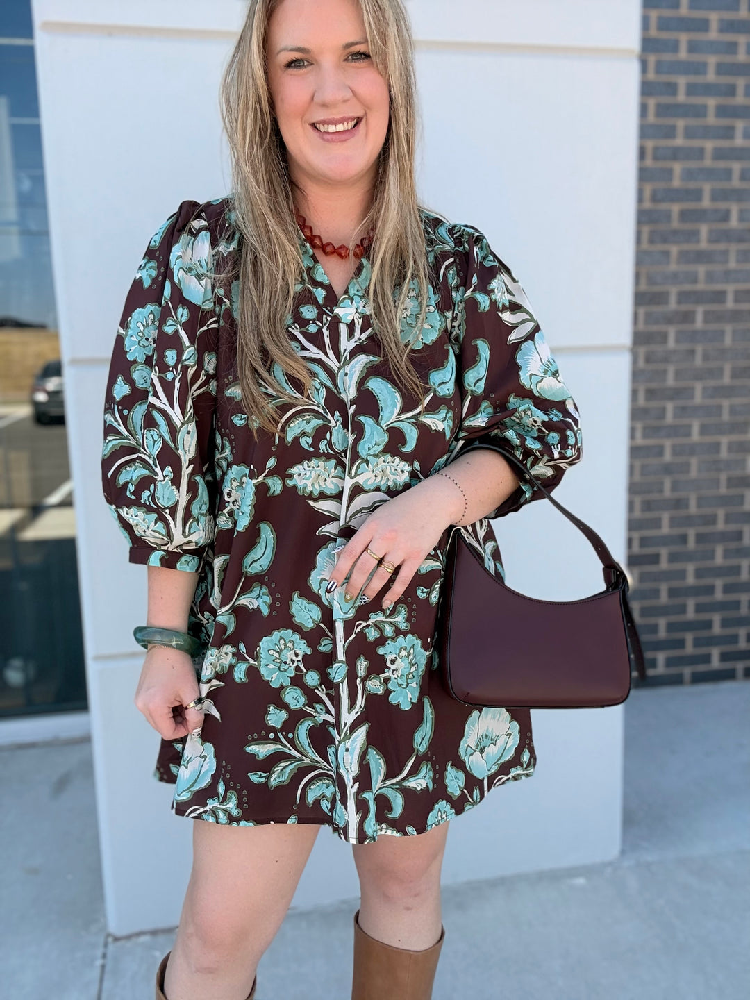 A woman wearing a brown puff-sleeve mini dress with a teal and white floral pattern, holding a dark plum shoulder bag, standing outdoors in front of a white wall and brick column.