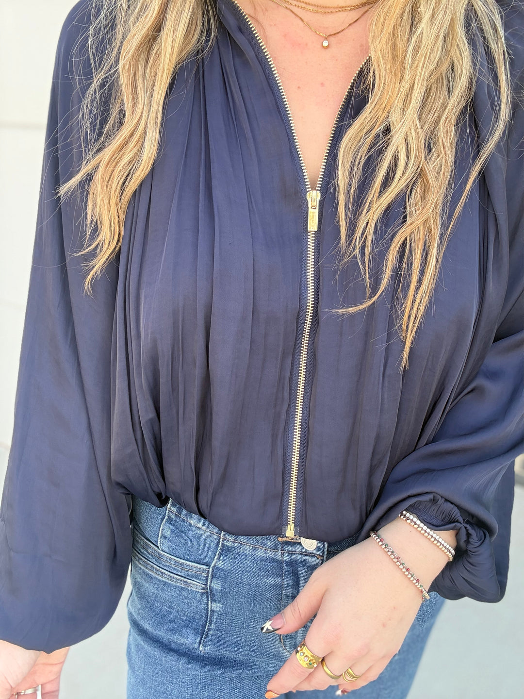 A woman wearing a navy blue long-sleeve zip-up jacket with a gold zipper, paired with blue jeans and gold jewelry.