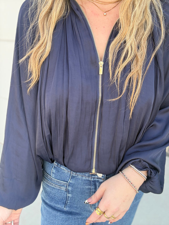A woman wearing a navy blue long-sleeve zip-up jacket with a gold zipper, paired with blue jeans and gold jewelry.
