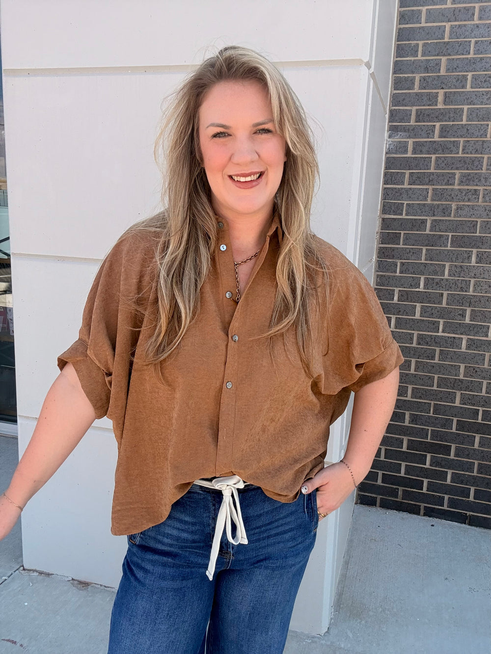 A woman wearing a brown short-sleeve button-up shirt with rolled sleeves and blue jeans, standing outdoors in front of a white wall and a dark brick column.