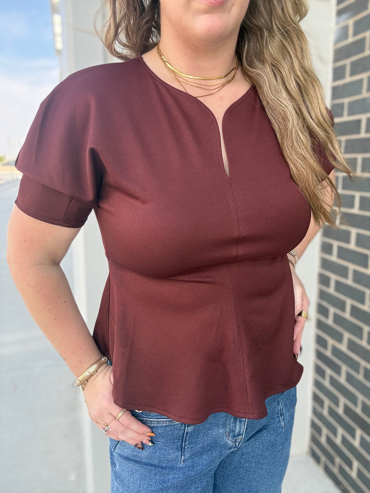 A woman wearing a short-sleeve maroon top with a split neckline and peplum hem, paired with blue jeans, standing outdoors next to a brick wall.