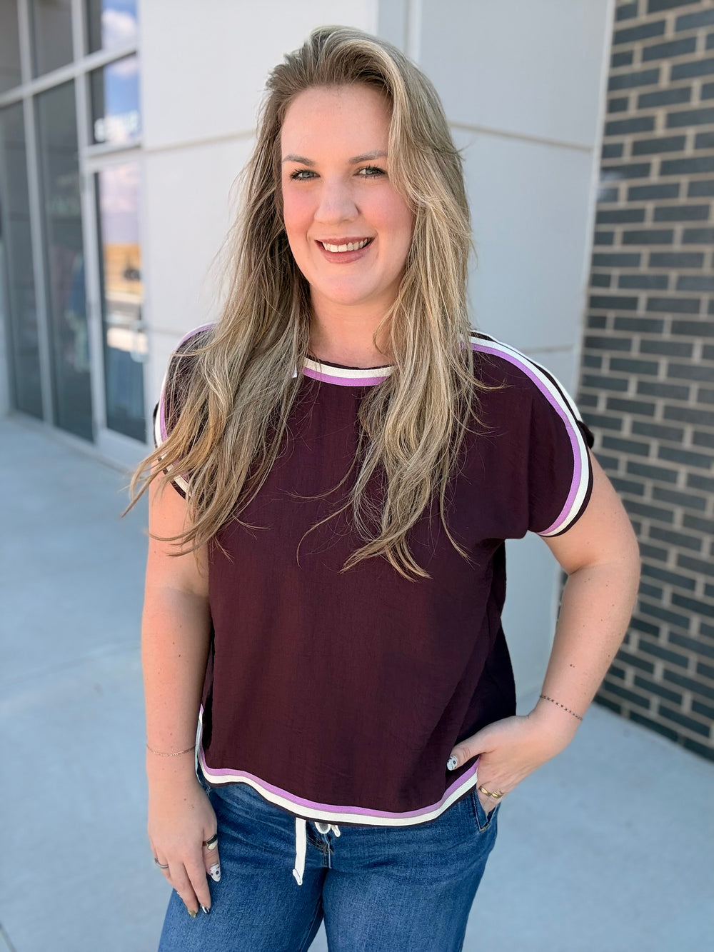 A woman with blonde hair wearing a short-sleeve maroon t-shirt with white and light purple trim, paired with blue jeans, standing outdoors in front of a modern building.