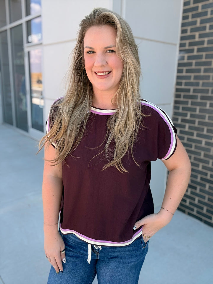A woman with blonde hair wearing a short-sleeve maroon t-shirt with white and light purple trim, paired with blue jeans, standing outdoors in front of a modern building.