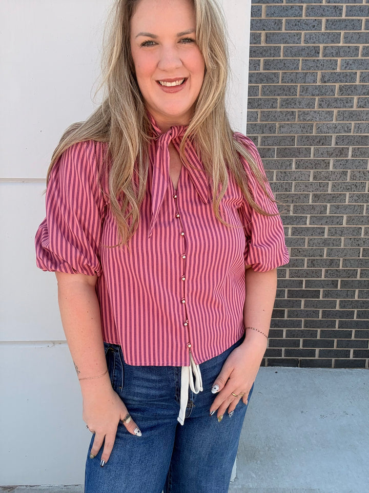 A woman wearing a pink and white vertically striped short-sleeve blouse with puff sleeves, a tie-neck detail, and front buttons, paired with blue jeans, standing outdoors against a white wall and a gray brick wall.