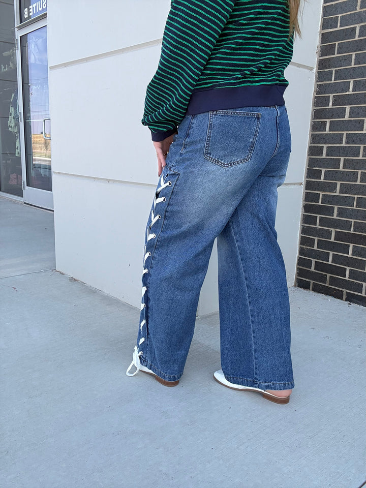 A person shown from the back wearing wide-leg blue jeans with white lace-up detailing down the side, a green and navy striped long-sleeve top, and white shoes, standing outdoors against a white building wall and brick column.