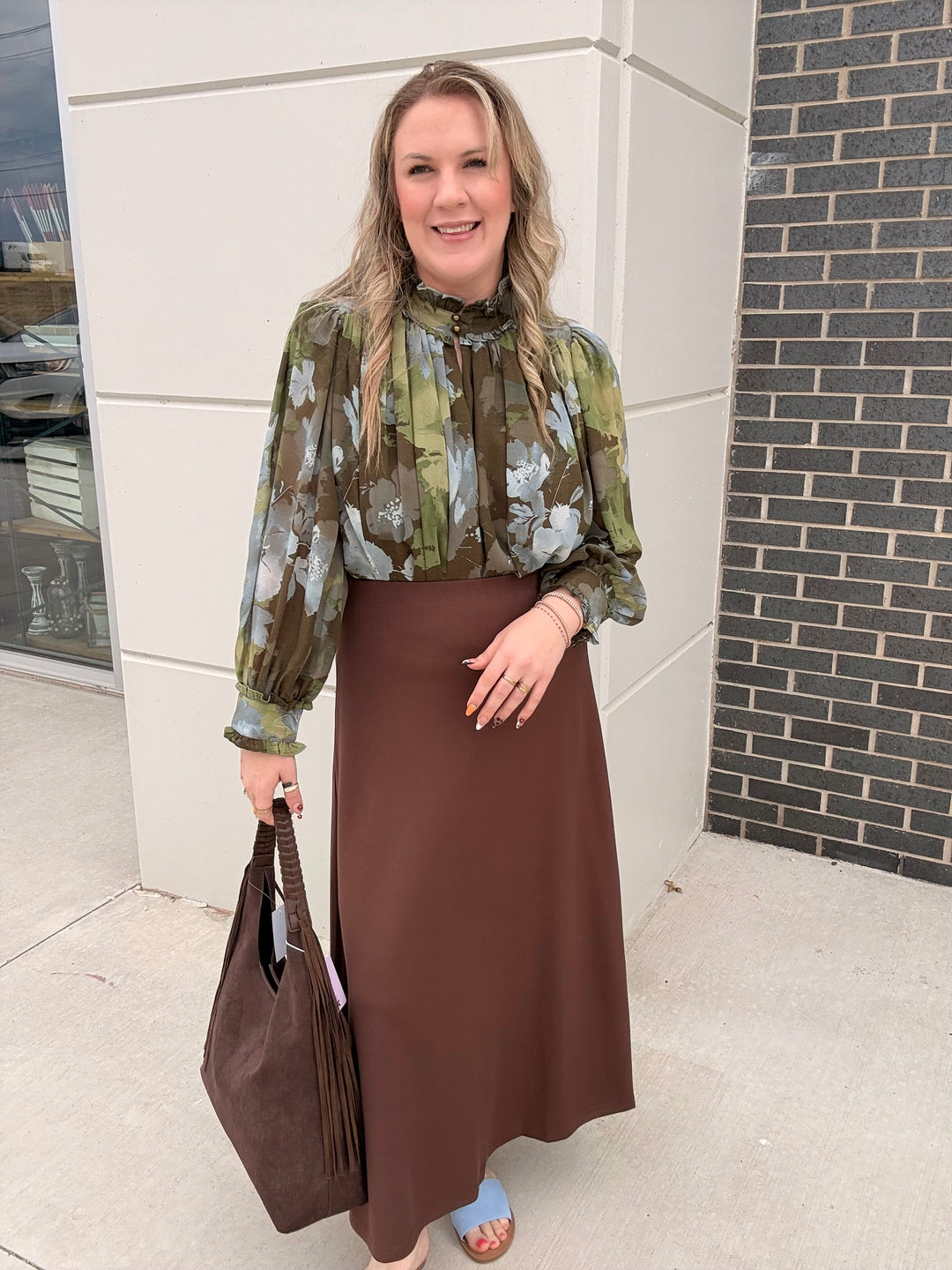 A woman stands outdoors wearing a long-sleeve high-neck blouse with an abstract green, olive, and gray pattern, paired with a solid brown midi skirt. She is holding a large brown tote bag.
