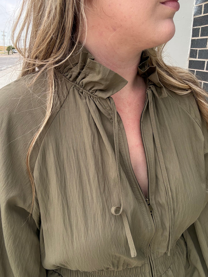 A close-up of a woman wearing an olive green lightweight zip-front jacket with a high gathered collar and drawstring details.
