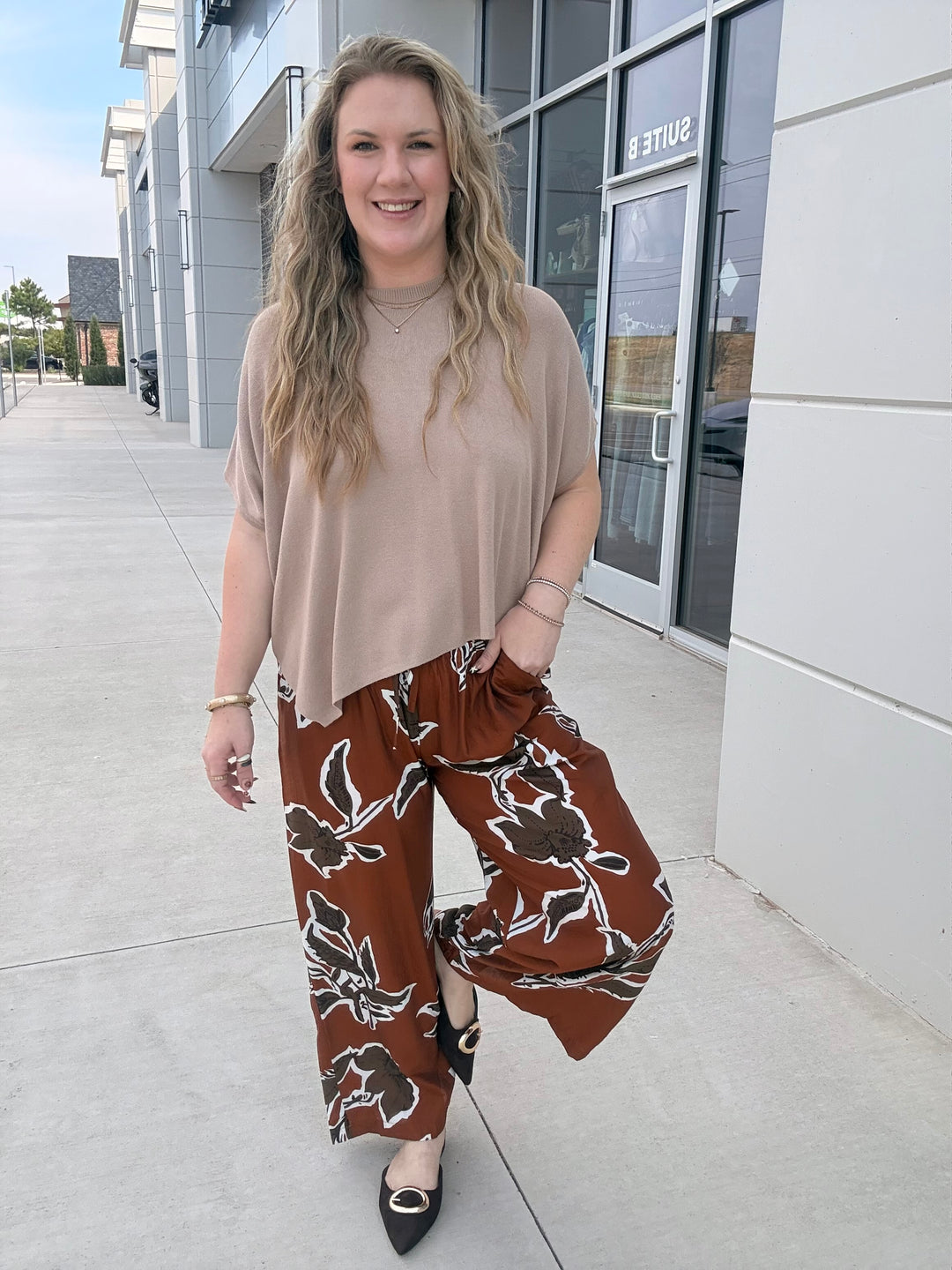 A woman stands outdoors on a sidewalk in front of a modern building, wearing a beige short-sleeve top and brown wide-leg pants with a white floral pattern.