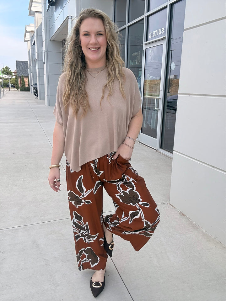 A woman stands outdoors on a sidewalk in front of a modern building, wearing a beige short-sleeve top and brown wide-leg pants with a white floral pattern.
