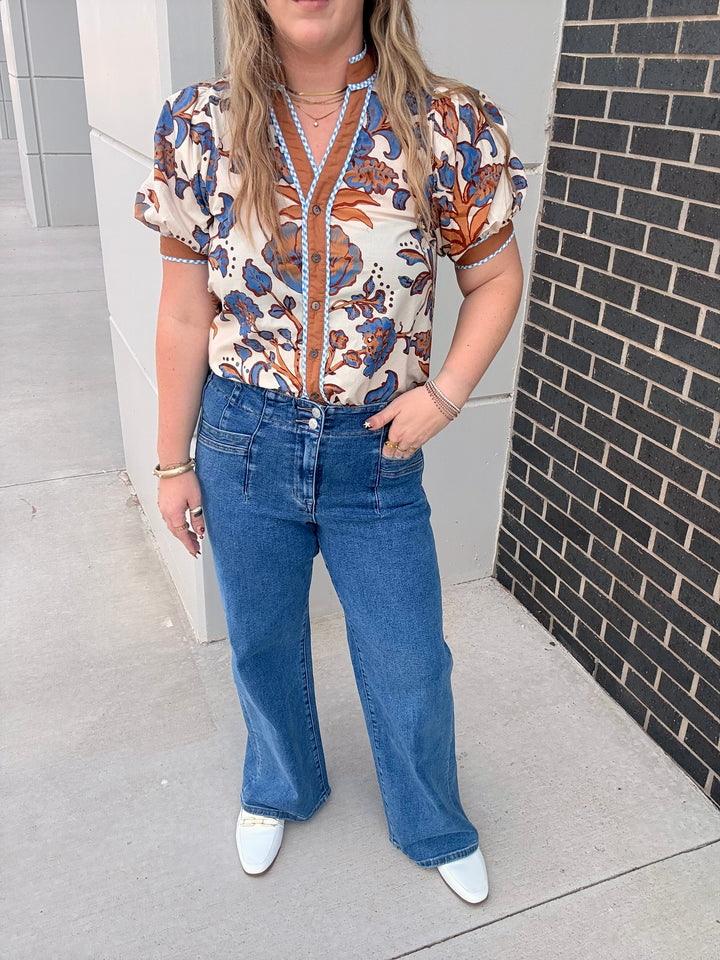 A woman wearing a short-sleeve button-front blouse with a cream base and orange, blue, and brown floral pattern, paired with wide-leg blue jeans and white pointed shoes, standing outdoors next to a gray wall and a black brick wall.