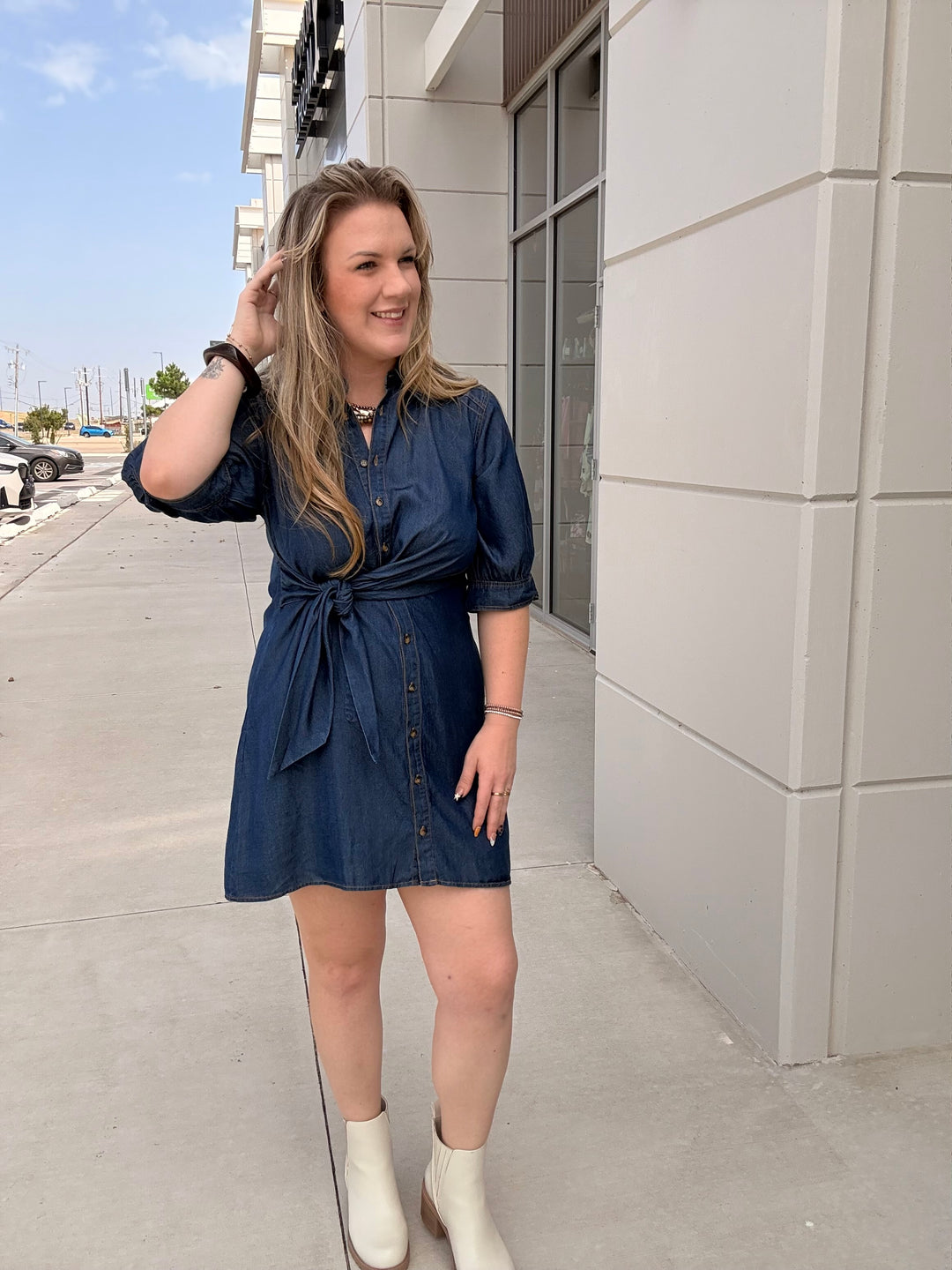 A woman wearing a dark blue denim-style mini dress with puff sleeves and a tie waist, standing outdoors on a sidewalk next to a modern building.