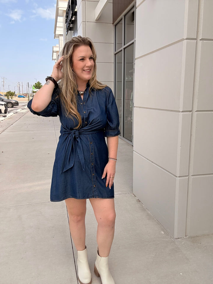 A woman wearing a dark blue denim-style mini dress with puff sleeves and a tie waist, standing outdoors on a sidewalk next to a modern building.