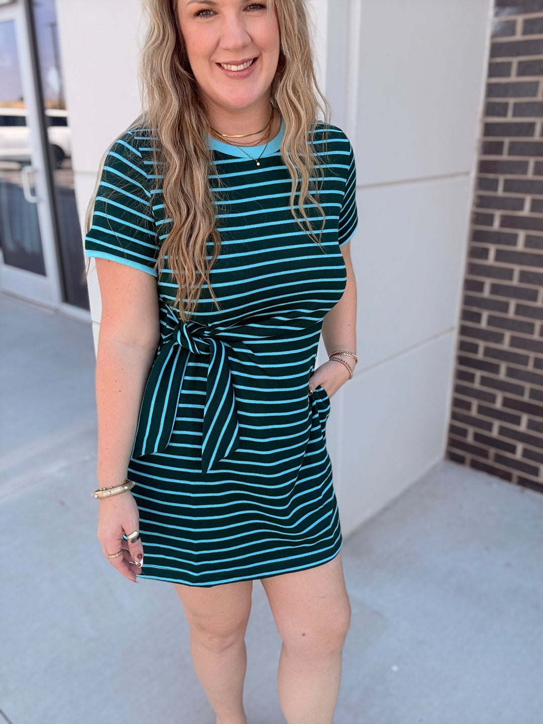 A woman wearing a short-sleeve mini dress with a dark teal and light blue horizontal stripe pattern, featuring a tie-waist detail and side pockets, standing outdoors.