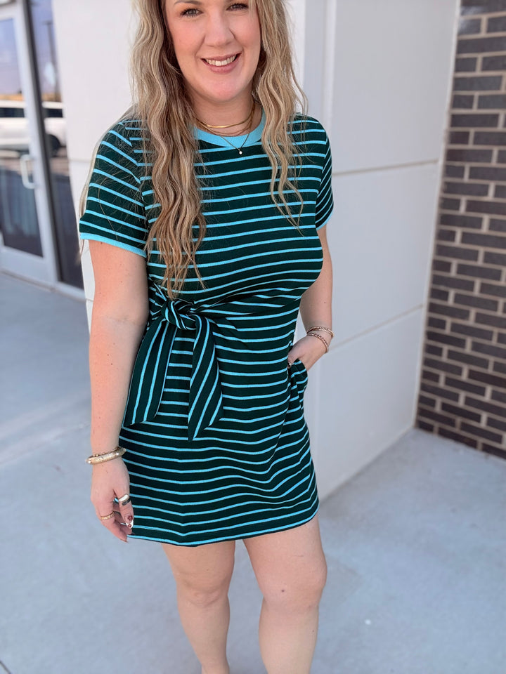 A woman wearing a short-sleeve mini dress with a dark teal and light blue horizontal stripe pattern, featuring a tie-waist detail and side pockets, standing outdoors.