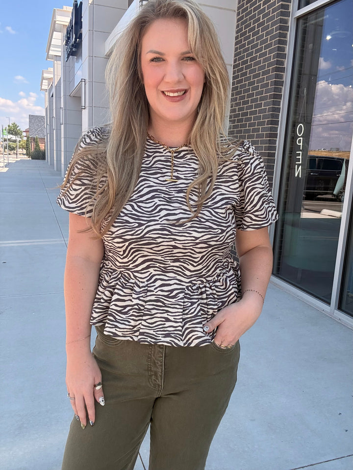 A woman wearing a short-sleeve zebra-print top and olive green pants, standing outdoors in front of a storefront.