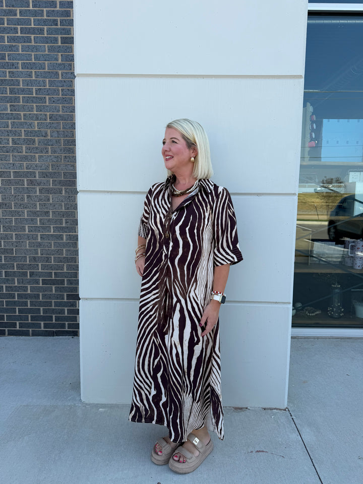 A woman with blonde hair stands outdoors in front of a white wall, wearing a long brown and white zebra-print shirt dress and beige platform sandals.