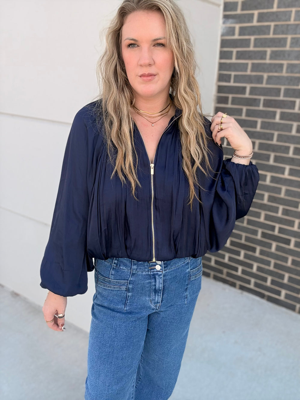 A woman wearing a navy blue long-sleeve blouse with a front zipper and blue jeans, standing outdoors against a white wall and a dark brick wall.