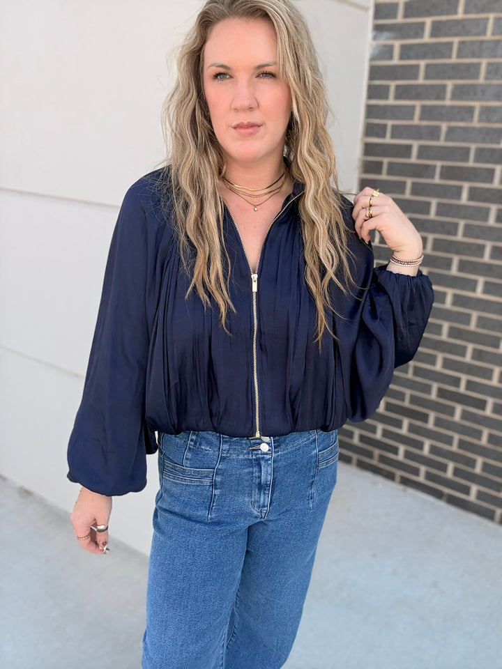 A woman wearing a navy blue long-sleeve blouse with a front zipper and blue jeans, standing outdoors against a white wall and a dark brick wall.