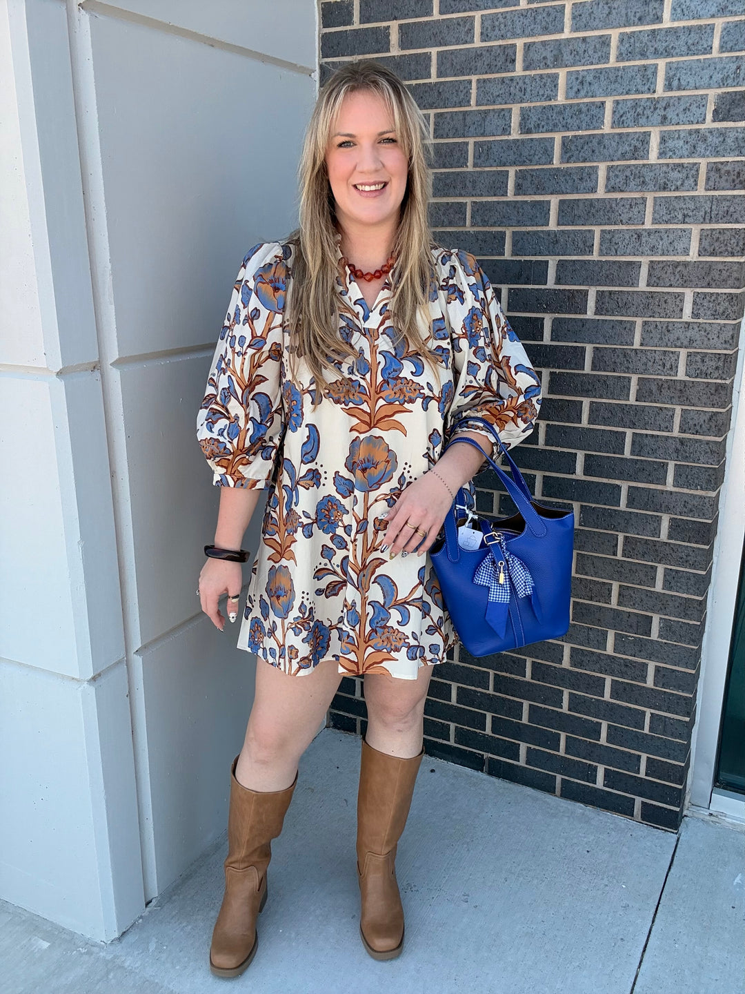 A woman stands outdoors against a gray brick wall and white column, wearing a short-sleeve bohemian-style mini dress with a blue, brown, and orange floral pattern on a cream background. She is wearing tan knee-high boots and carrying a bright blue tote bag.