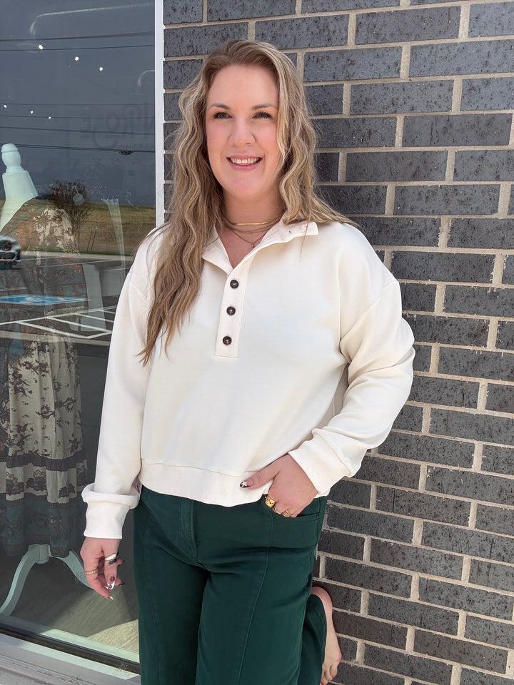 A woman with blonde hair stands in front of a gray brick wall wearing a cream-colored long-sleeve henley sweatshirt and dark green pants.