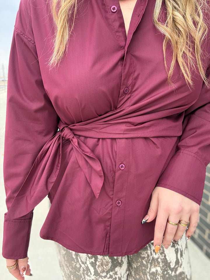 A woman wearing a long-sleeve wrap-style blouse in a deep berry/plum color, tied at the waist, with a button front and patterned gray pants.