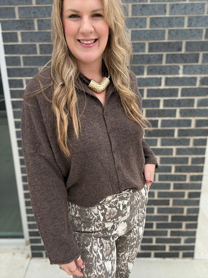 A woman wearing a brown long-sleeve zip-front top and camouflage-patterned pants, standing in front of a gray brick wall.