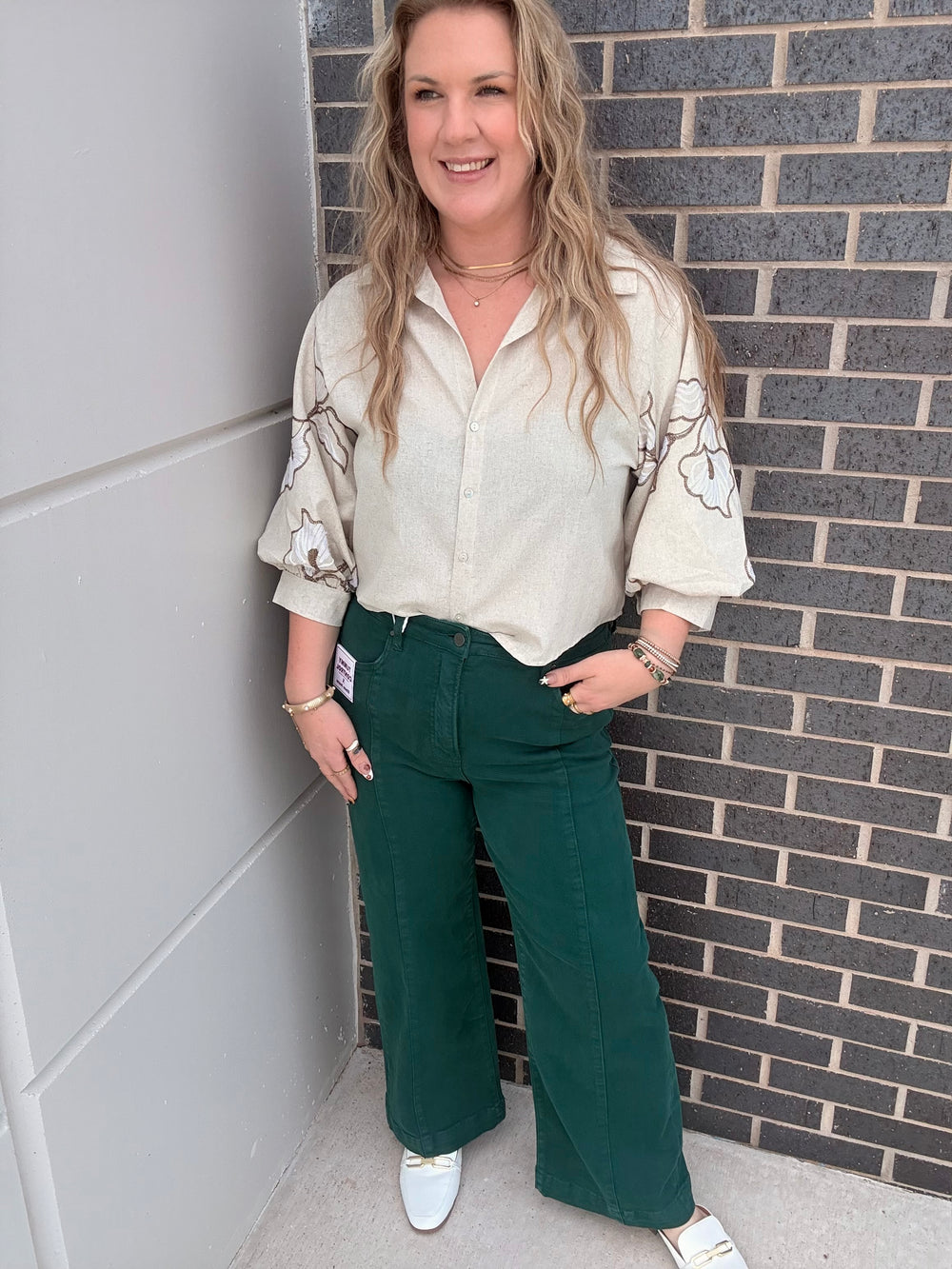 A woman stands outdoors against a gray brick wall and white door, wearing a cream button-up shirt with embroidered details on the sleeves and dark green wide-leg pants.