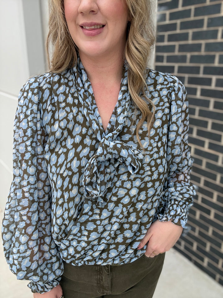 A woman wearing a light blue and olive green leopard-print blouse with a tie-neck detail, paired with olive green pants, standing in front of a brick wall.