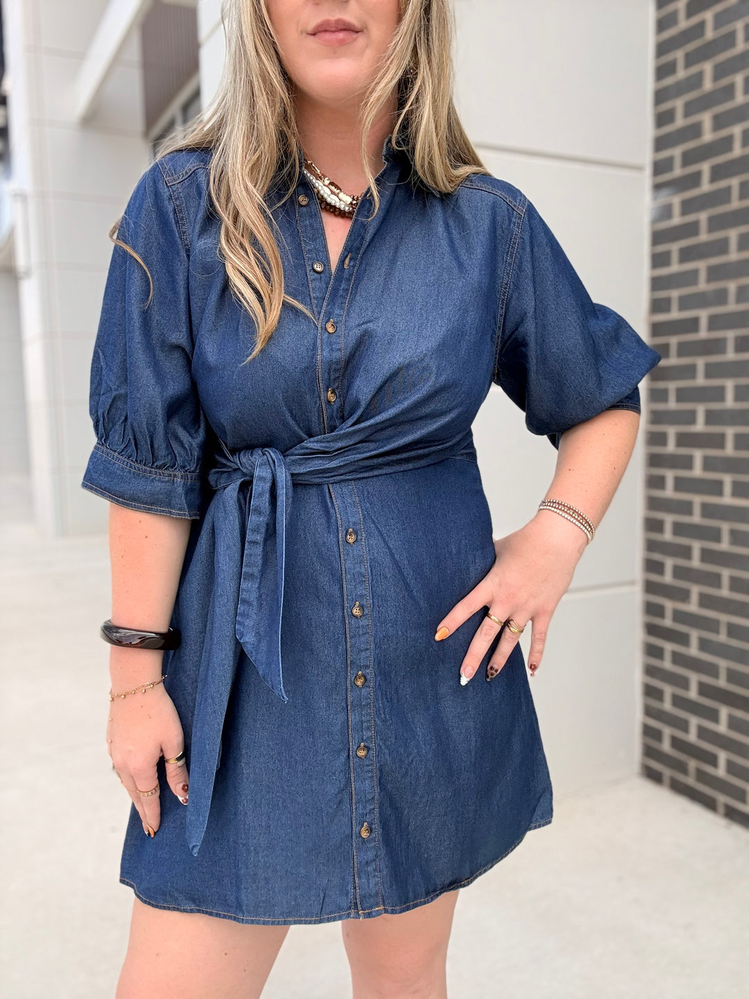 A woman wearing a blue denim mini dress with puff sleeves, a button-down front, and a self-tie waist, photographed outdoors against a modern building wall.
