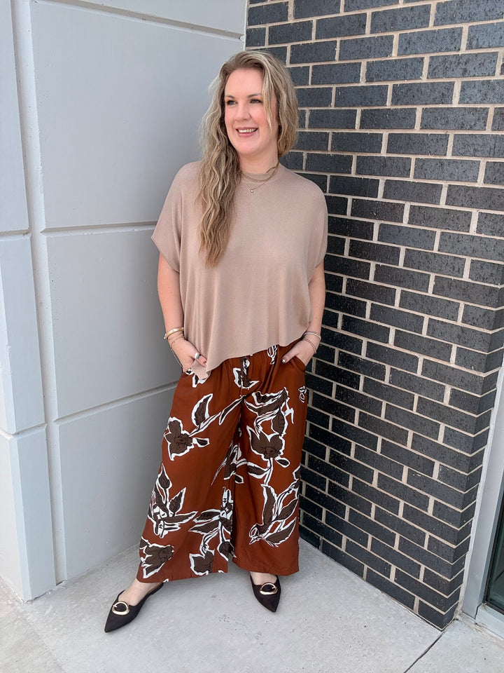 A woman stands outdoors against a gray paneled wall and dark brick wall, wearing a beige short-sleeve mock-neck top and brown wide-leg pants with a white floral pattern.