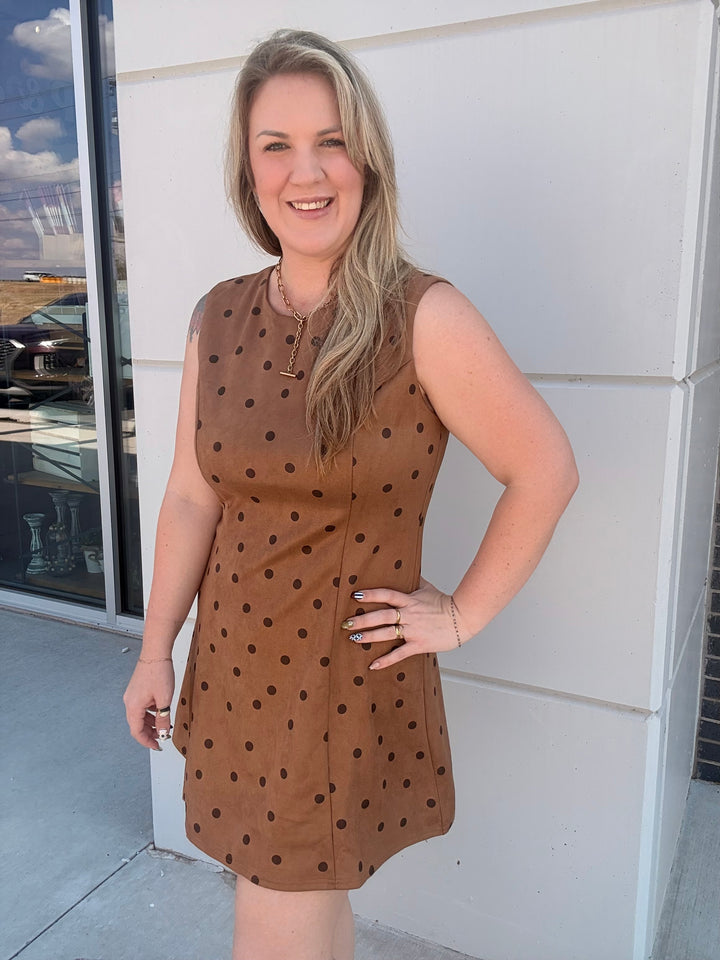 A woman modeling a sleeveless brown dress with black polka dots, standing outdoors against a white building wall.