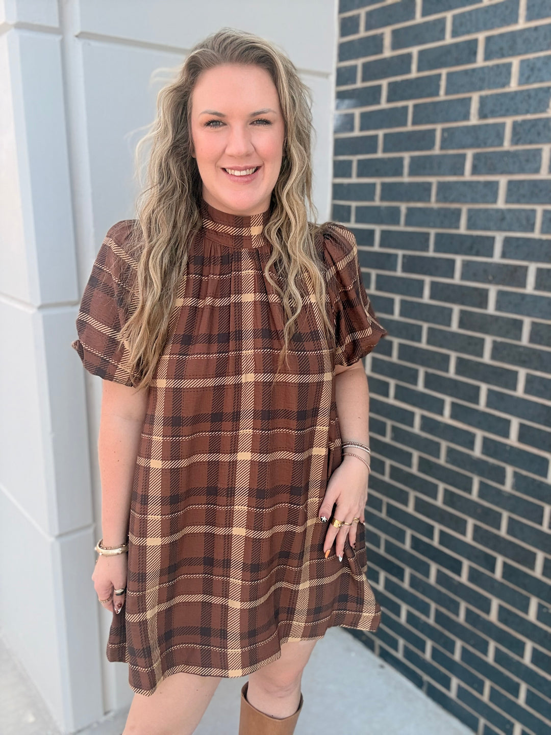 A woman wearing a brown and beige plaid short-sleeve dress, standing outdoors against a gray brick wall and white building corner.
