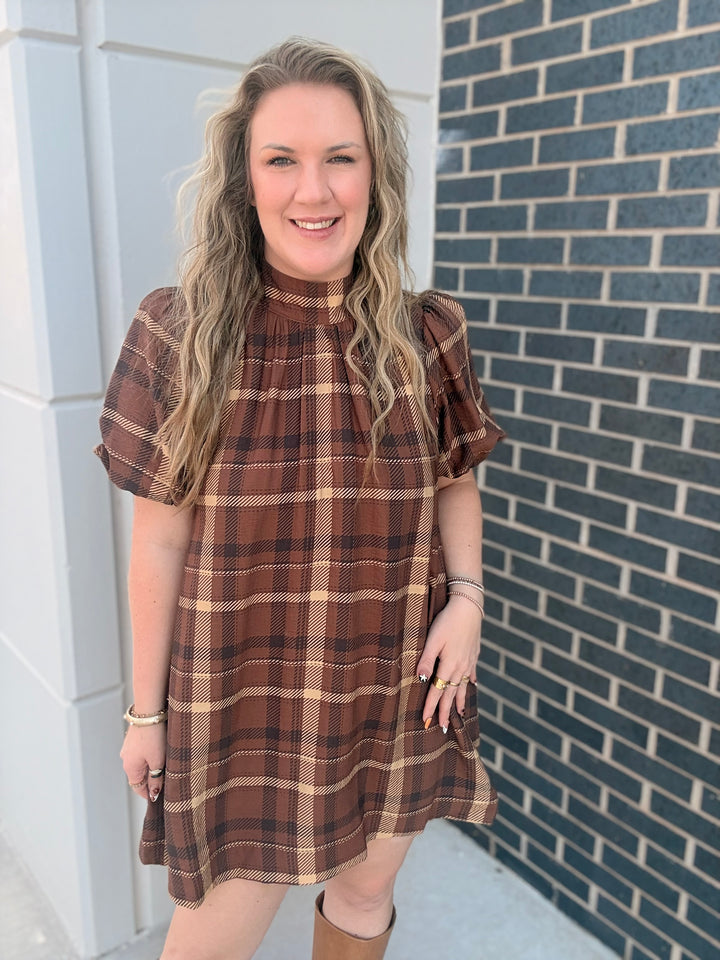 A woman wearing a brown and beige plaid short-sleeve dress, standing outdoors against a gray brick wall and white building corner.