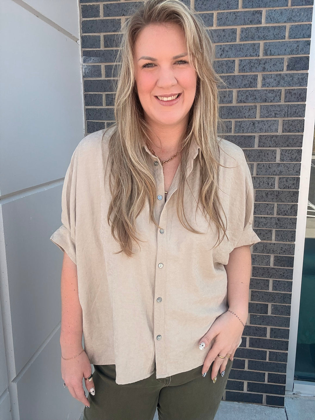A smiling blonde woman wearing a light beige short-sleeve linen button-up shirt and olive green pants, standing outdoors in front of a gray brick wall.