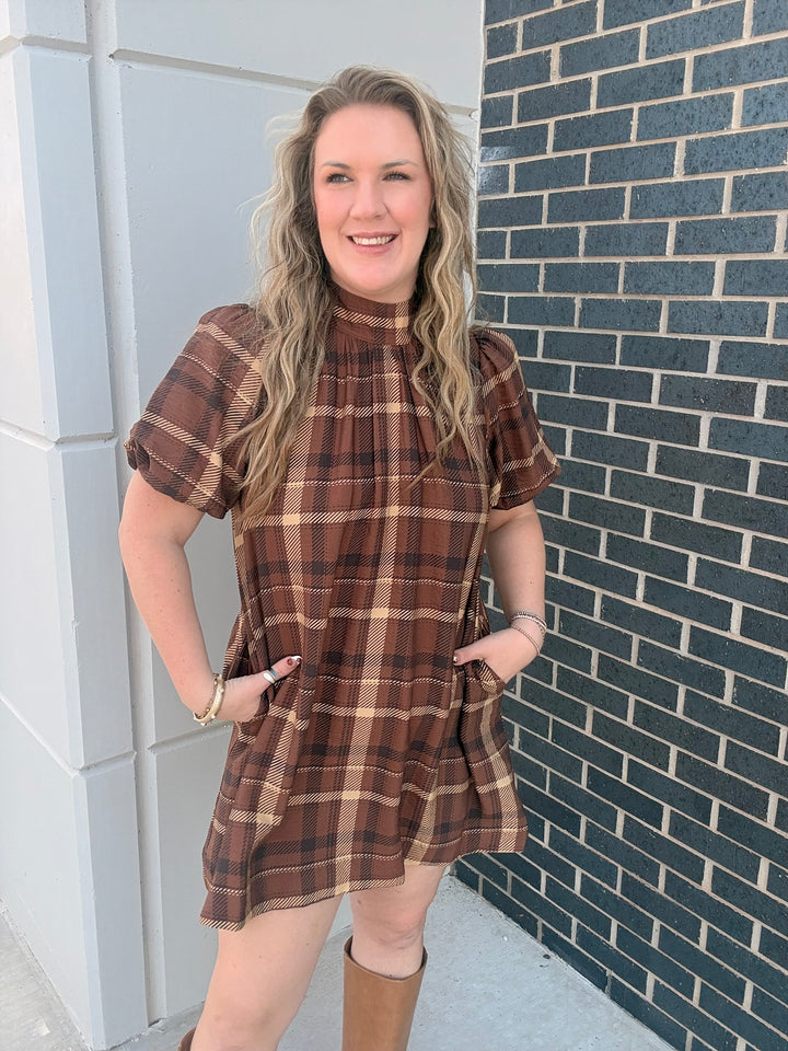 A woman wearing a brown and beige plaid short-sleeve mini dress with pockets, standing outdoors against a gray brick wall and white tiled wall.