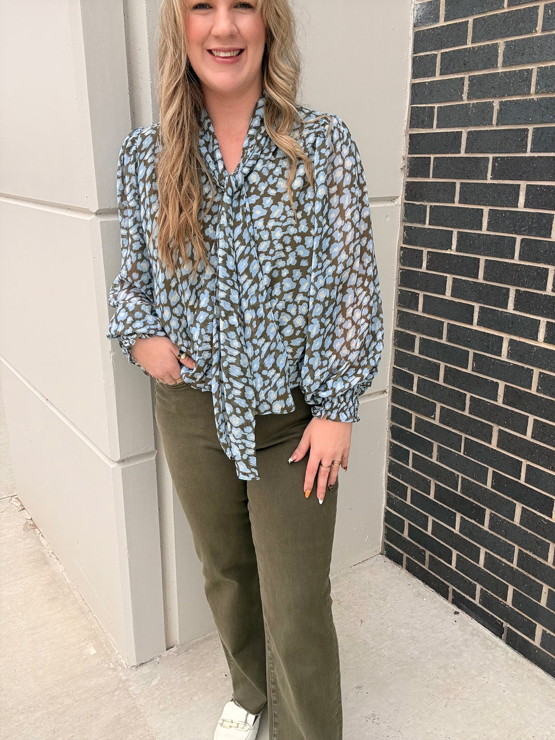 A woman wearing a light blue and brown leopard print blouse with long balloon sleeves, tucked into olive green wide-leg pants, standing outdoors next to a gray wall and a black brick wall.