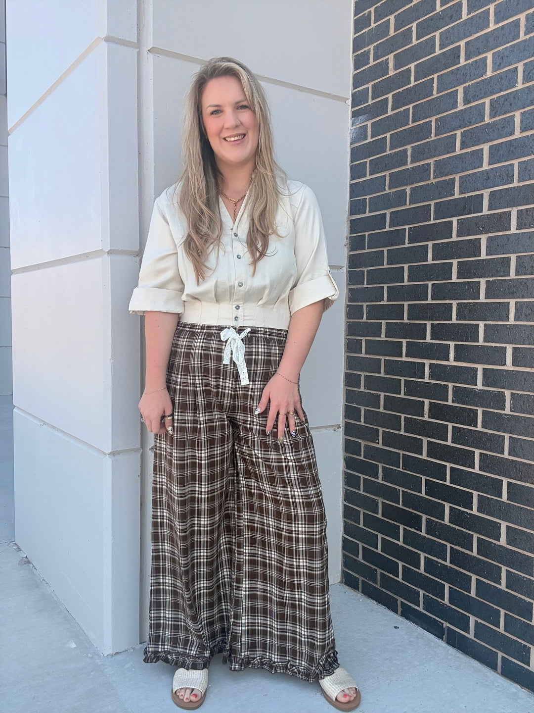 A woman stands outdoors against a white concrete pillar and a dark brick wall, wearing a cream short-sleeve button-up shirt and brown plaid wide-leg pants with a white tie at the waist.