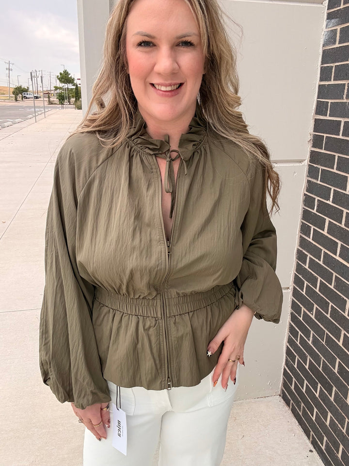 A woman wearing an olive green zip-up jacket with a tie-neck detail and elastic waist, paired with white pants, standing outdoors next to a brick wall.