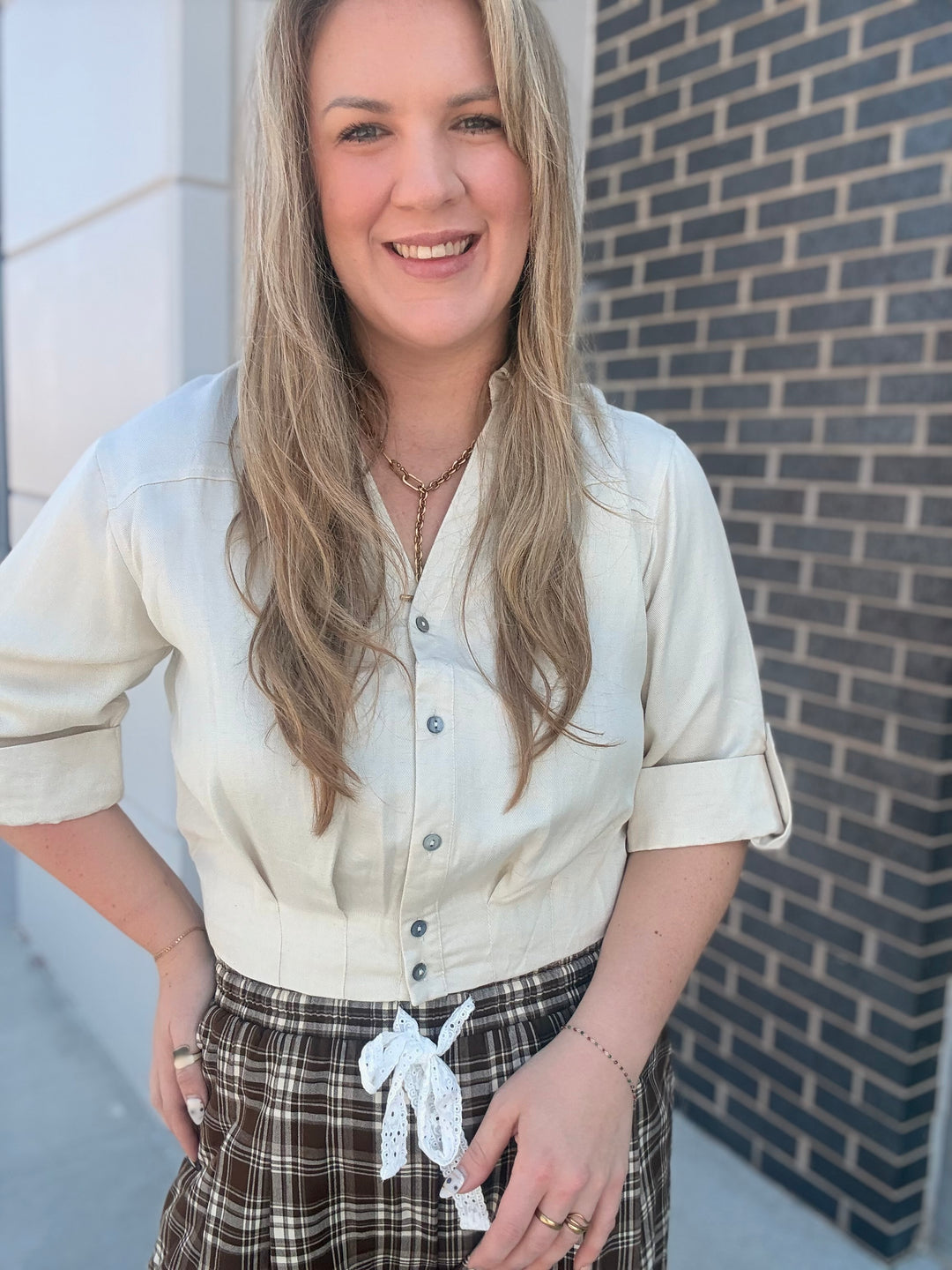 A woman with long blonde hair smiling, wearing a cream-colored short-sleeve button-up shirt and a brown plaid skirt with a white lace tie, standing outdoors against a brick wall.