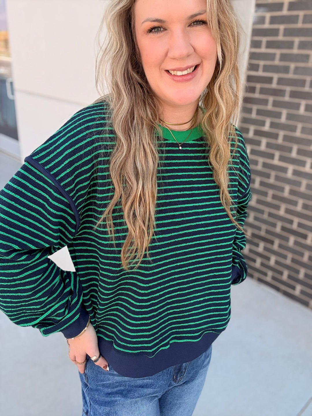 A woman wearing a green and navy striped long-sleeve knit top with balloon sleeves, paired with blue jeans, standing outdoors against a brick wall.