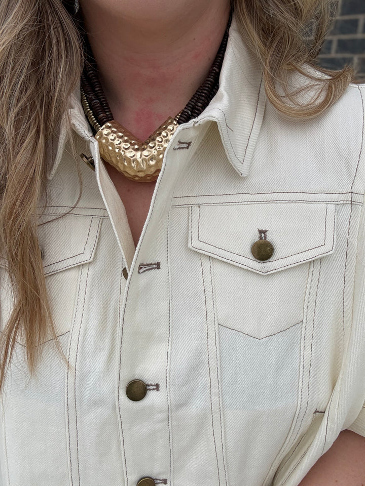 Close-up of a person wearing a cream-colored denim shirt-jacket with brass buttons and a chunky gold statement necklace.