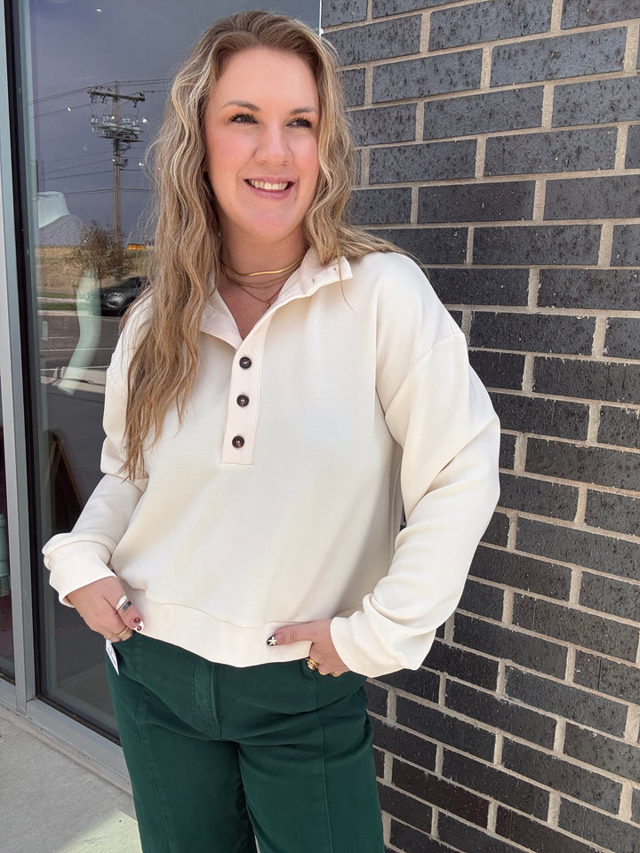 A woman wearing a cream-colored long-sleeve henley sweatshirt and dark green pants, standing outdoors against a dark gray brick wall.