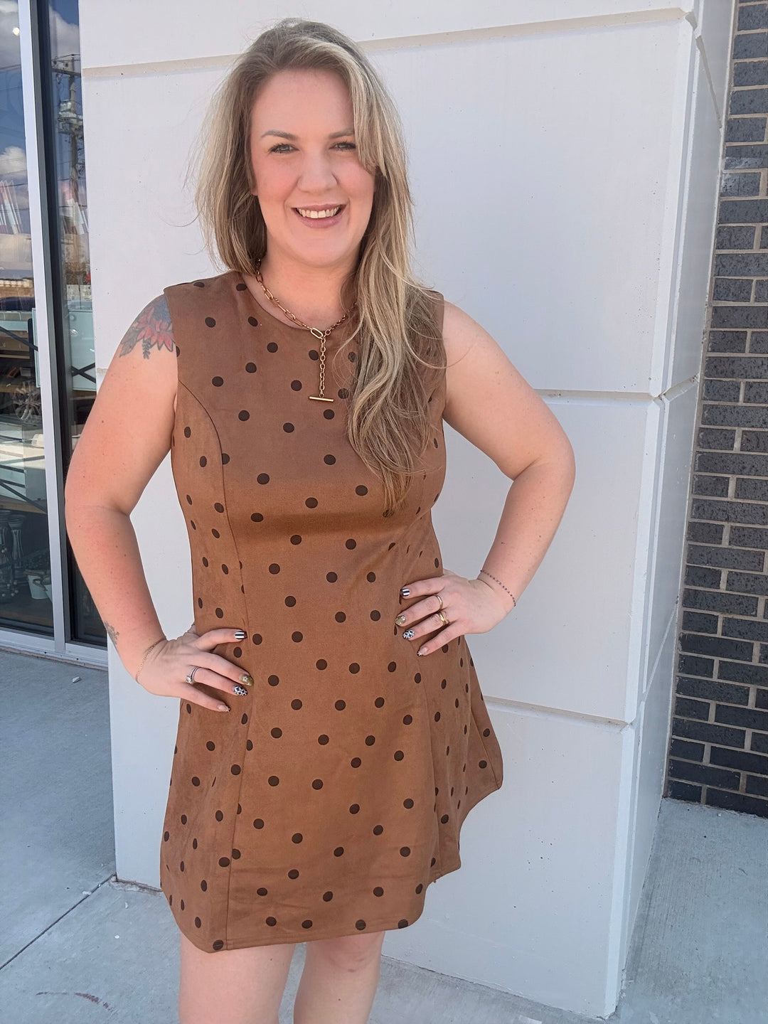 A woman wearing a sleeveless brown dress with black polka dots, standing outdoors in front of a white building wall.