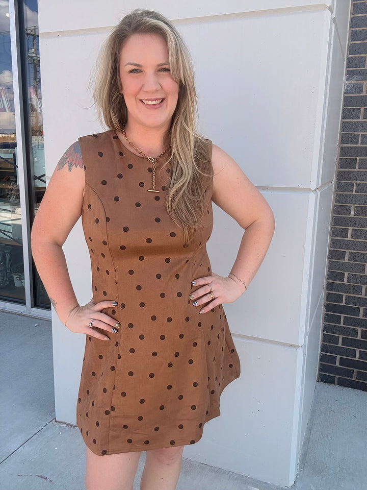 A woman wearing a sleeveless brown dress with black polka dots, standing outdoors in front of a white building wall.