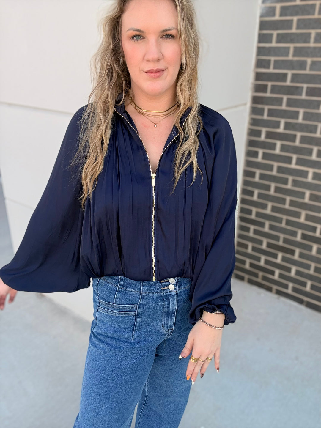 A woman wearing a navy blue long-sleeve blouse with a front zipper and blue jeans, standing outdoors against a light gray wall and brick column.