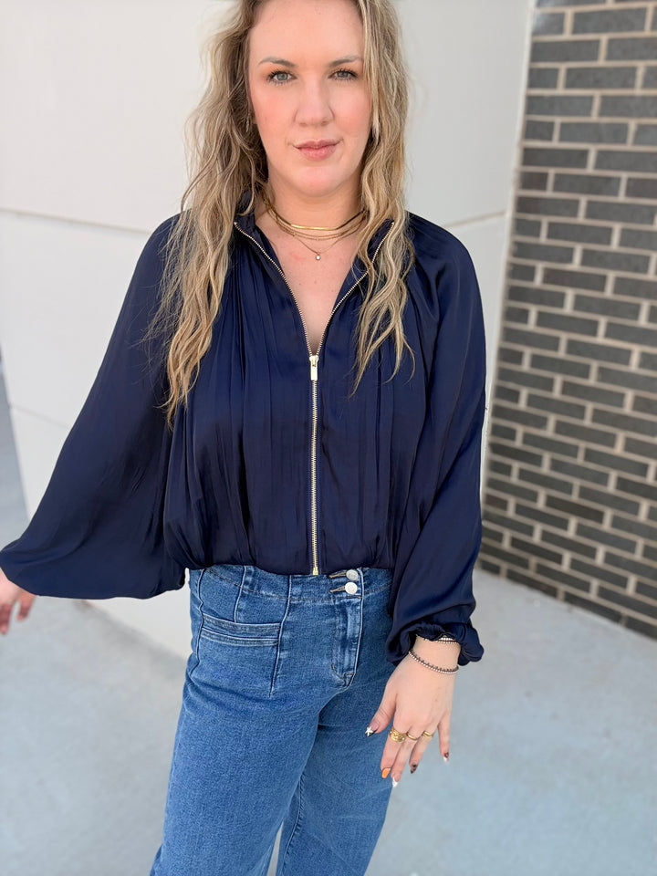 A woman wearing a navy blue long-sleeve blouse with a front zipper and blue jeans, standing outdoors against a light gray wall and brick column.